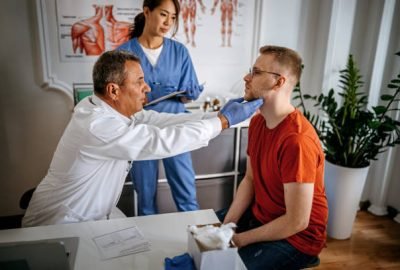 Doctor examining a young man patient in his medical office while nurse writing information about patient