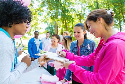 Diverse group of adults and teenagers are lined up at registration table in sunny park. They are signing up to run in charity 5k race or marathon to raise money for breast cancer research. People are wearing pink athletic clothing and breast cancer awareness ribbons.