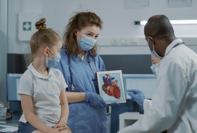 Woman nurse showing cardiology image on digital tablet, letting doctor explain cardiovascular diagnosis to child and parent. Assistant holding modern device with heart anatomy picture.